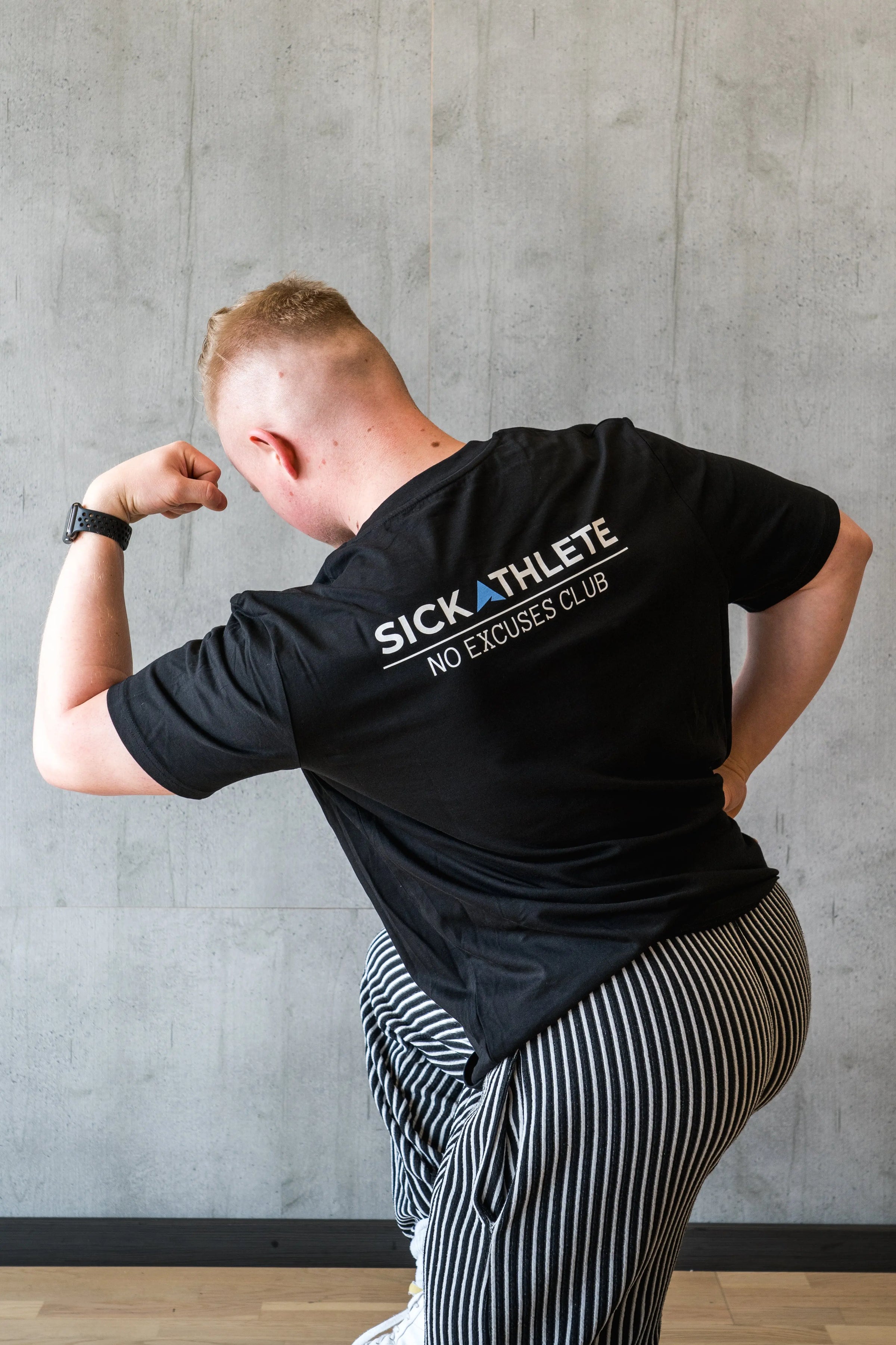 Bodybuilder im Gym der bei einer Pose seinen Rücken und den Bizeps flext, dabei ist das SICKATHLETE NO EXCUSES CLUB LOGO im Vordergrund