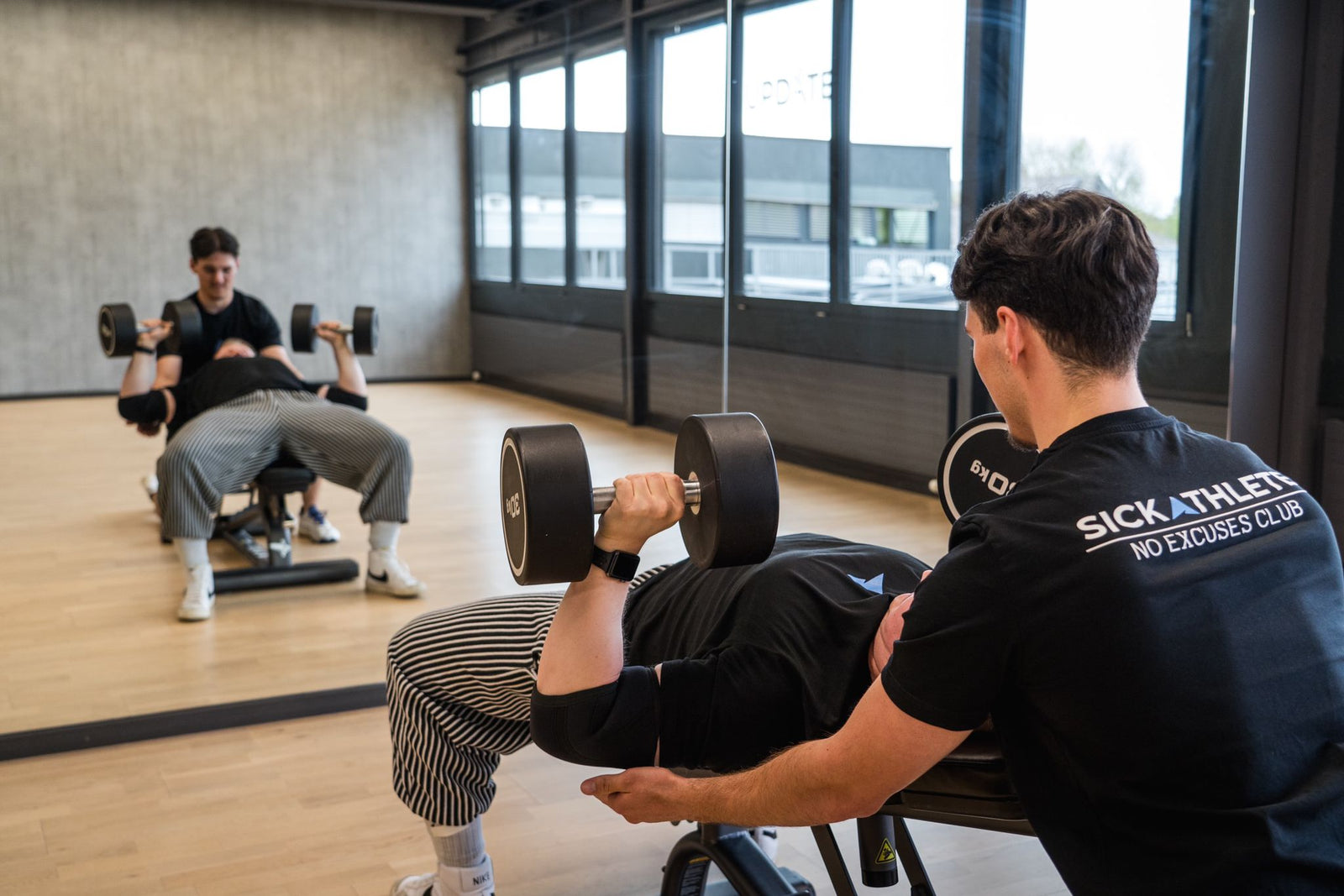 Zwei Sportler unterstützen sich im Gym beim Bankdrücken und tragen ein SICKATHLETE T-Shirt