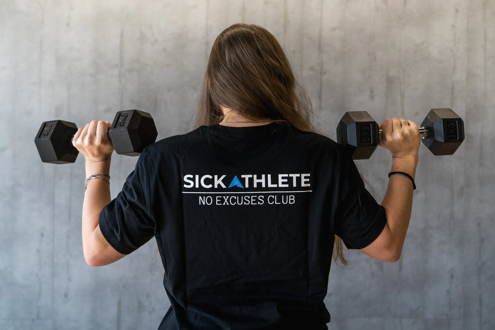 Sportlerin mit SICKATHLETE NO EXCUSES Club T-Shirt trägt während Training im Gym zwei Hanteln