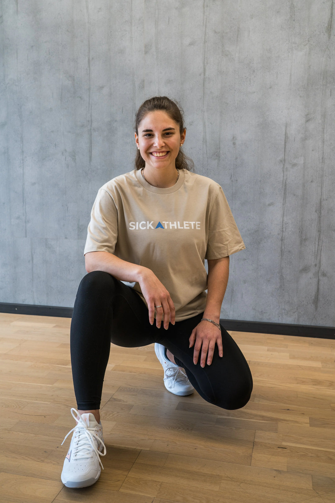 Fröhliche Sportlerin kniend vor einer grauen Wand trägt das SICKATHLETE ESSENTIAL T-Shirt