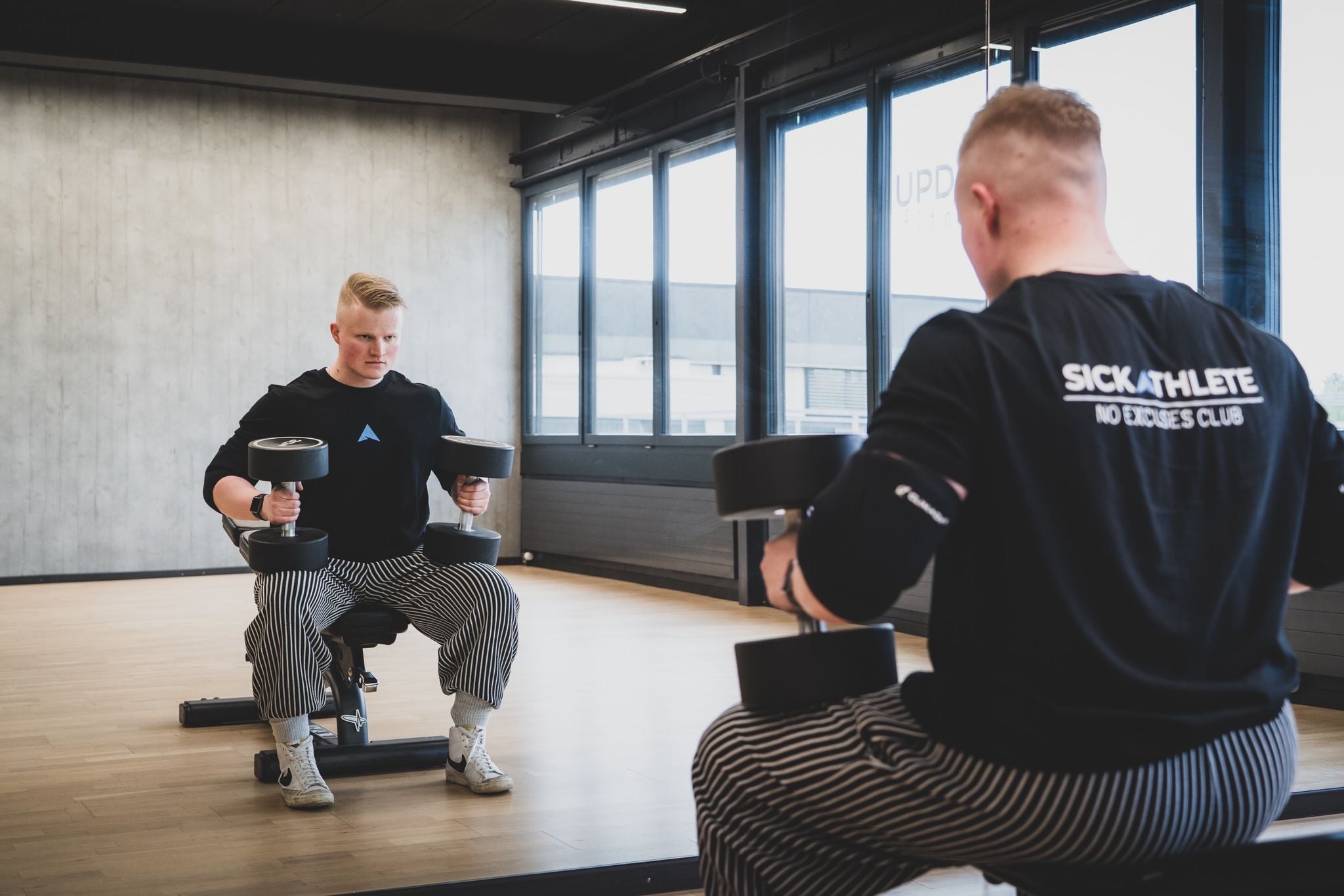 Mann im Gym mit zwei Kurzhanteln der ein SICKATHLETE Club T-Shirt trägt