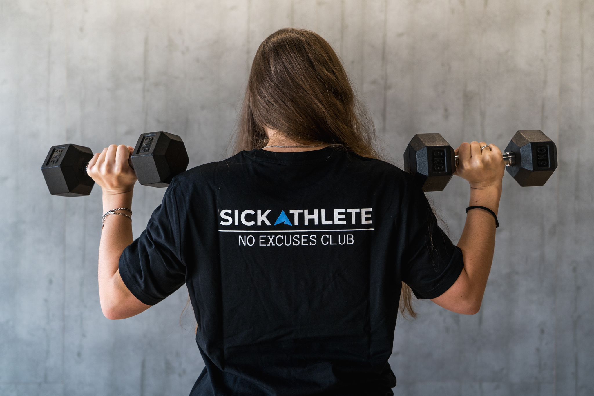 Sportlerin mit SICKATHLETE NO EXCUSES Club T-Shirt trägt während Training im Gym zwei Hanteln