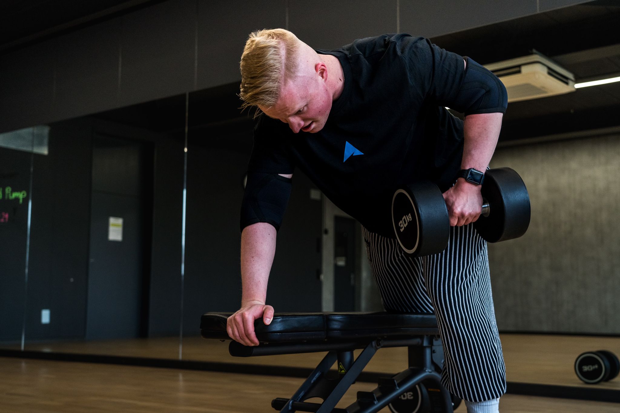 Junger Athlet der mit einem SICKATHLETE Club T-Shirt kniend auf einer Hantelbank den Rücken trainiert